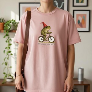 May include: A light pink t-shirt featuring a whimsical illustration of a green frog riding a bicycle and wearing a red hat. The text "Off to cause a kerfuffle" is printed below the image. The shirt is made of soft material.