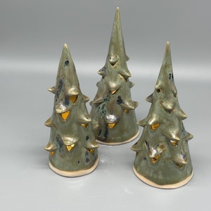 May include: Three ceramic Christmas tree ornaments. The trees are glazed in shades of green with gold accents, and have a textured, layered design. The trees are arranged on a neutral grey surface, creating a festive holiday display.