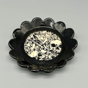 May include: Black ceramic dish with a scalloped edge. The dish has a white center with a black and white design of skulls and bones.