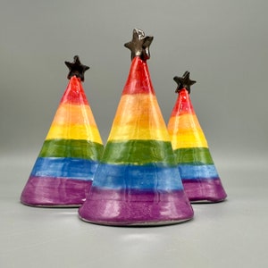 Può includere: Tre sculture coniche in ceramica con una gamma di colori arcobaleno, sormontate da una stella scura. I colori vanno dal rosso al viola, con strisce gialle, verdi e blu. Le sculture sono su sfondo grigio.