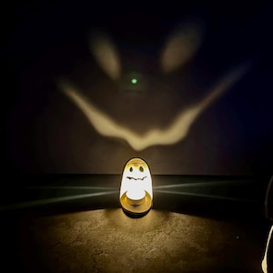 Può includere: Una lampada cilindrica bianca con un design a forma di fantasma ritagliato. La sorgente luminosa interna proietta una grande ombra spettrale e luminosa di un fantasma con le braccia tese sulla parete dietro l'oggetto in una stanza buia.