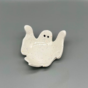 Può includere: Un piatto in ceramica bianca a forma di fantasma con occhi neri e una glassa macchiettata. Il piatto ha la forma di un fantasma con le braccia distese e una testa rotonda.