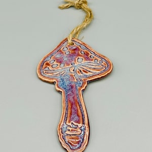 Könnte beinhalten: Ein Keramik-Pilz-Anhänger mit brauner und violetter Glasur. Der Anhänger hat eine Schnur zum Aufhängen.