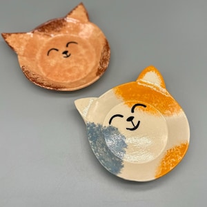 Może przedstawiać: Dwa ceramiczne talerze w kształcie kota. Jeden jest pomarańczowy z brązowymi krawędziami, drugi biały, pomarańczowy i niebieski. Oba mają uśmiechniętą twarz w kolorze czarnym. Te dekoracyjne talerze są idealne do przechowywania drobnych przedmiotów.