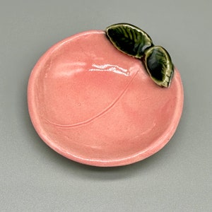 May include: A small pink ceramic dish shaped like a peach with two green leaves on top.