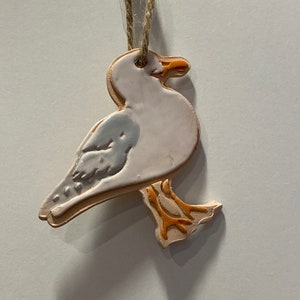 Pode incluir: Um ornamento de cerâmica branca em forma de gaivota com pés e bico laranja. O ornamento está pendurado em uma corda.