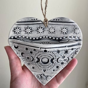 Könnte beinhalten: Weißer, herzförmiger Wandbehang aus Keramik mit einem schwarz-weißen Design aus Sternen und Monden. Das Herz hat oben eine kleine Tasche, in der kleine Gegenstände aufbewahrt werden können.