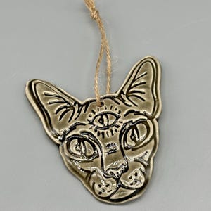 May include: A ceramic cat face ornament with a third eye design. The ornament is a muted green color with black detailing and a jute string for hanging. The cat's features are stylized with etched lines and a glossy finish.