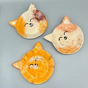 Puede incluir: Tres platos de cerámica hechos a mano con forma de gato. Cada plato presenta una cara sonriente y está pintado en tonos naranja, marrón y crema. Los platos tienen un diseño único y caprichoso, perfecto para servir aperitivos o como piezas decorativas.