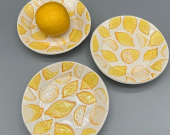Könnte beinhalten: Drei weiße Keramikplatten mit Zitronenmotiv. Jede Platte ist mit gelben Zitronenscheiben und ganzen Zitronen verziert. Eine Platte enthält eine ganze Zitrone. Die Teller sind auf einer grauen Oberfläche angeordnet.
