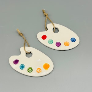 May include: Two off-white ceramic artist palette ornaments with colorful paint splotches and a jute string for hanging. Each palette has a hole for the string and is decorated with red, green, blue, purple, and orange paint.