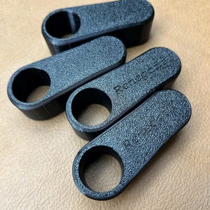 Pu&ograve; includere: Quattro apribottiglie neri, strutturati, stampati in 3D. Ogni apribottiglie ha un'apertura circolare su un'estremit&agrave; e la scritta "Renegade" sulla parte superiore. Gli apribottiglie sono disposti su una superficie marrone.
