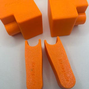 May include: Four bright orange 3D-printed objects are displayed against a white background. Two larger, angular pieces with the word "Renegade" are visible, along with two smaller, curved pieces also bearing the "Renegade" text. The objects have a matte finish.