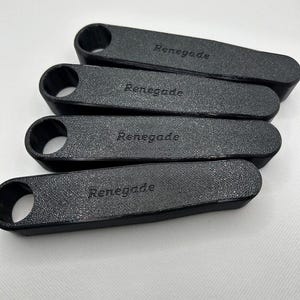 Pu&ograve; includere: Quattro oggetti rettangolari neri e strutturati con un foro arrotondato su un'estremit&agrave;. La parola "Renegade" &egrave; stampata su ogni oggetto. Gli oggetti sono disposti in modo leggermente sovrapposto su uno sfondo bianco.