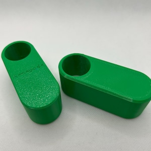 May include: Two bright green, rectangular 3D-printed objects. Each has a cylindrical hole on one end. The objects are on a white surface. One object has the text "omega.de" printed on it.