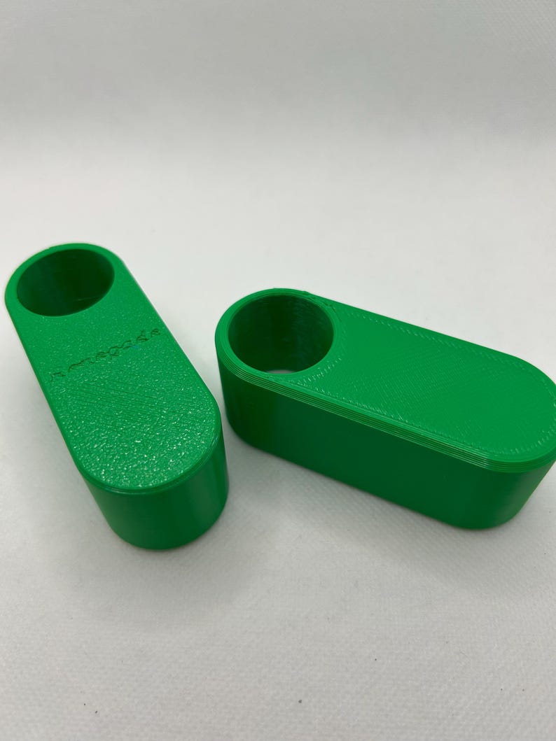 May include: Two bright green, rectangular 3D-printed objects. Each has a cylindrical hole on one end. The objects are on a white surface. One object has the text "omega.de" printed on it.