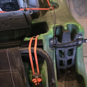May include: Close-up of a green kayak with black and orange accents. The kayak features a black seat frame, orange cords, and a gray storage compartment. The kayak is designed for water activities.