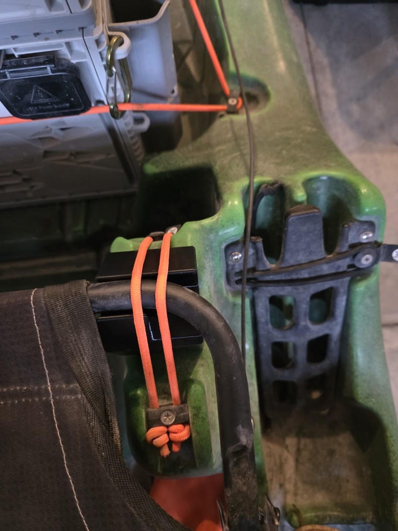 May include: Close-up of a green kayak with black and orange accents. The kayak features a black seat frame, orange cords, and a gray storage compartment. The kayak is designed for water activities.