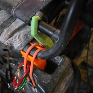 May include: Close-up of climbing gear, featuring a black carabiner, orange rope, and a green plastic component. The gear is attached to a black metal bar and a gray and black fabric.