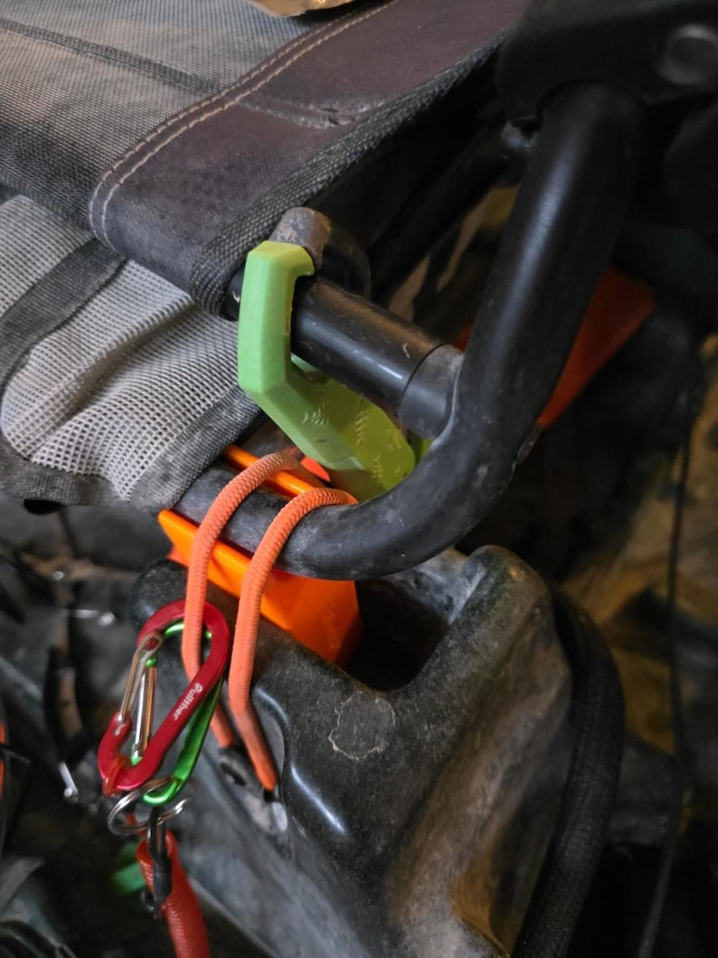 May include: Close-up of climbing gear, featuring a black carabiner, orange rope, and a green plastic component. The gear is attached to a black metal bar and a gray and black fabric.