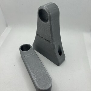 May include: Two gray 3D-printed objects. One is a tall, angled piece with a circular hole at the top and a smaller hole near the bottom. The other is a rectangular piece with a circular hole. Both pieces have the word "Renegade" printed on them.