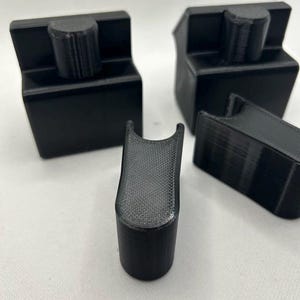 May include: Four black 3D-printed objects are displayed against a white background. Two are rectangular with a cylindrical top, one is a U-shaped piece, and the last is a rectangular piece. The objects appear to be components of a larger item.