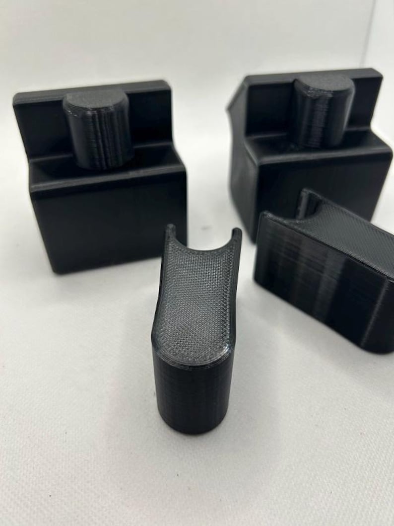 May include: Four black 3D-printed objects are displayed against a white background. Two are rectangular with a cylindrical top, one is a U-shaped piece, and the last is a rectangular piece. The objects appear to be components of a larger item.