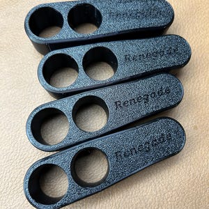 Pu&ograve; includere: Quattro oggetti in plastica nera, strutturati, ciascuno con due fori circolari. La parola "Renegade" &egrave; in rilievo su ciascuno. Sono impilati su uno sfondo marrone chiaro. Gli oggetti sembrano essere stampati in 3D.