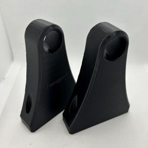 May include: Two black, 3D-printed brackets with a curved top and a circular hole. Each bracket has a smaller hole near the base. The brackets are designed with a textured surface and are set against a white background.