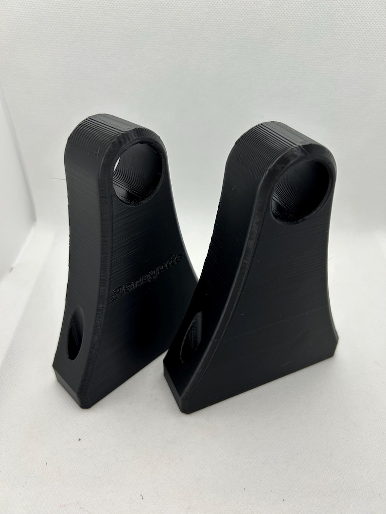 May include: Two black, 3D-printed brackets with a curved top and a circular hole. Each bracket has a smaller hole near the base. The brackets are designed with a textured surface and are set against a white background.