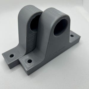 Puede incluir: Dos soportes grises impresos en 3D. Cada soporte tiene una parte superior curva con una abertura circular y una base rectangular con dos agujeros para el montaje. La palabra "Renegade" est&aacute; impresa en el lateral de un soporte.