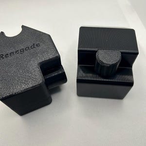 May include: Two black, geometric-shaped objects. One has the word "Renegade" embossed on its surface. The other object has a cylindrical knob. Both objects appear to be made of a textured, matte material. The objects are set against a plain white background.