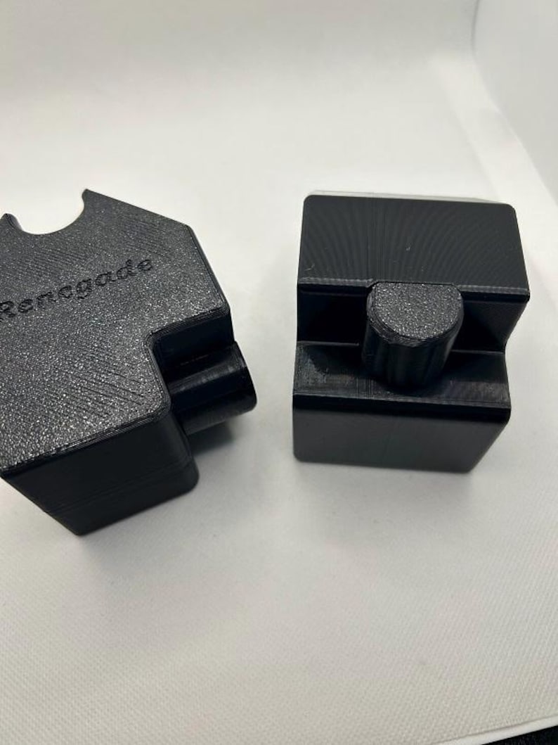 May include: Two black, geometric-shaped objects. One has the word "Renegade" embossed on its surface. The other object has a cylindrical knob. Both objects appear to be made of a textured, matte material. The objects are set against a plain white background.