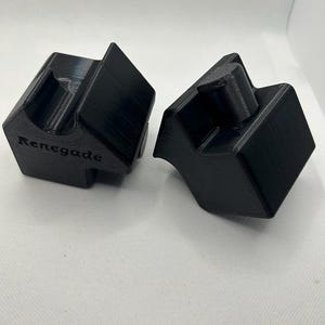 May include: Two black, geometric, 3D-printed objects. Each has a unique shape with angled surfaces and a central, raised cylindrical feature. The word "Renegade" is printed on one of the objects. The objects are displayed on a white surface.