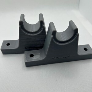 Puede incluir: Dos soportes grises impresos en 3D con un dise&ntilde;o de cuna curvada y una base plana con un agujero para el montaje. La palabra "Renegade" est&aacute; impresa en un soporte.