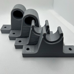 Puede incluir: Tres soportes de tuber&iacute;a grises impresos en 3D. Cada soporte tiene una cuna curva y una base plana con orificios de montaje. La palabra "Renegade" est&aacute; impresa en la base de los soportes. Los soportes est&aacute;n dispuestos en fila.