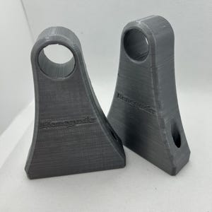 May include: Two gray 3D-printed brackets with a flared base and a circular opening at the top. The word "Renegade" is printed on the front of each bracket. The brackets are made of a textured material.