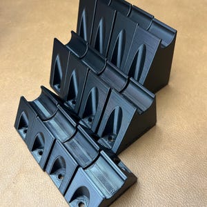 May include: Several black 3D-printed organizers are arranged on a brown surface. Each organizer has a curved top, a series of arched openings, and a flat base with holes. The organizers are angled, creating a dynamic display.