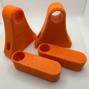 May include: Four bright orange 3D-printed objects. Two are tall with a circular opening at the top and a smaller hole on the side. The other two are rectangular with a circular opening. The word "Renegade" is printed on each object.