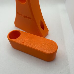 May include: Two bright orange 3D-printed objects. One is a tall, angled piece with two holes and the word "Renegade" embossed on it. The other is a smaller, rectangular piece with a single hole. Both have a textured surface.