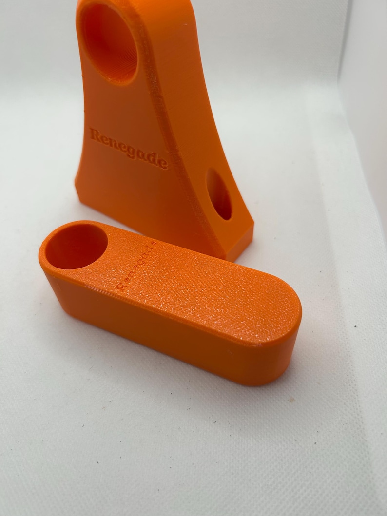 May include: Two bright orange 3D-printed objects. One is a tall, angled piece with two holes and the word "Renegade" embossed on it. The other is a smaller, rectangular piece with a single hole. Both have a textured surface.