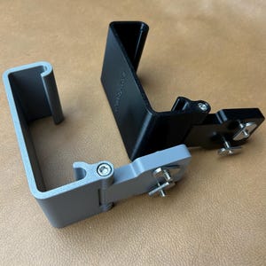 May include: Three metal brackets in gray and black. The gray bracket has a curved top and a hinged section with a screw. The black bracket is rectangular and has a hinged section with a screw and a wing nut.