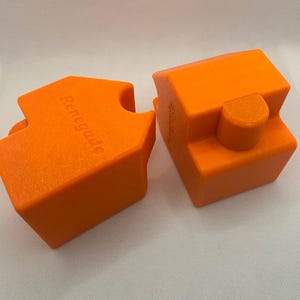 May include: Two bright orange, geometric blocks with the word "Renegade" embossed on one. The blocks have a textured surface and are designed to fit together. The background is a plain white.