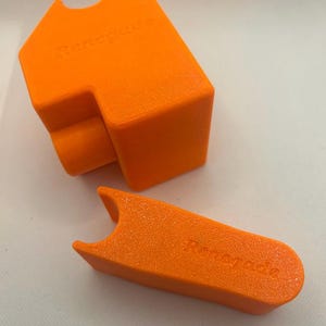 May include: Two bright orange, 3D-printed objects. One is a rectangular shape with a curved cutout and the word "Renegade" embossed on it. The other is a T-shaped piece with the word "Renegade" embossed on it.