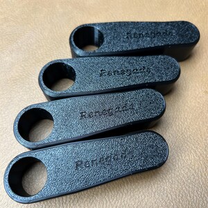 Pu&ograve; includere: Quattro oggetti neri, strutturati, di forma oblunga, con un'apertura circolare su un'estremit&agrave;. La parola "Renegade" &egrave; in rilievo su ogni oggetto. Gli oggetti sono disposti diagonalmente su una superficie beige.