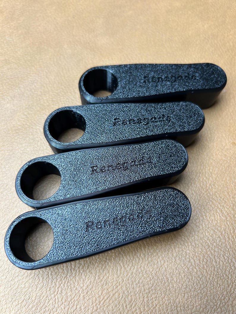 Pu&ograve; includere: Quattro oggetti neri, strutturati, di forma oblunga, con un'apertura circolare su un'estremit&agrave;. La parola "Renegade" &egrave; in rilievo su ogni oggetto. Gli oggetti sono disposti diagonalmente su una superficie beige.