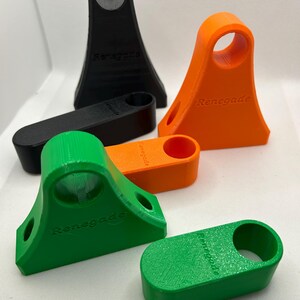 May include: A collection of 3D-printed Renegade phone stands in black, orange, and green. The stands have a distinctive design with a circular opening at the top and the word "Renegade" printed on the front.