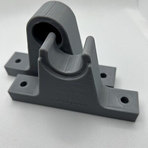 Puede incluir: Dos soportes grises impresos en 3D. Cada soporte tiene una base con dos agujeros para el montaje y una secci&oacute;n curva dise&ntilde;ada para sujetar un objeto cil&iacute;ndrico. El texto "renegade" est&aacute; impreso en la base de un soporte.