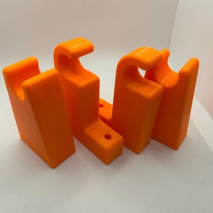 May include: Four bright orange, 3D-printed brackets. Each bracket has a curved top and a rectangular base with a hole. The brackets are arranged in a row, with the curved tops facing the same direction. The brackets are made of plastic.
