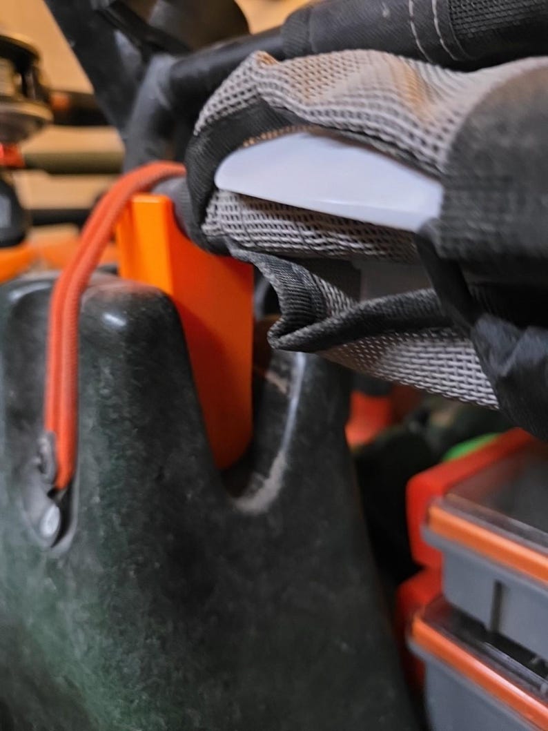 May include: Close-up of a black and orange kayak or boat component. The image shows a white handle, a mesh strap, and an orange plastic piece. A red rope is also visible. The components appear to be part of a larger system.